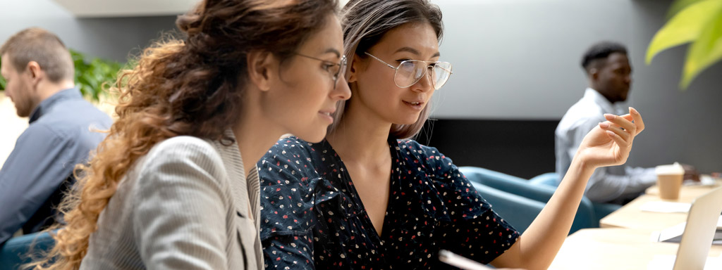 Two business woman looking at a laptop screen with other people in the background.